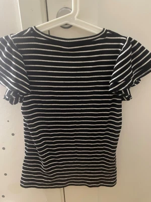 Randig svartvit topp med volangärm - Snygg topp med svarta och vita ränder och volangärmar. Ribbad struktur som ger en cool känsla och passar perfekt till jeans eller kjol. Rund halsringning och figurnära passform. Perfekt för dig som gillar en trendig och enkel stil.