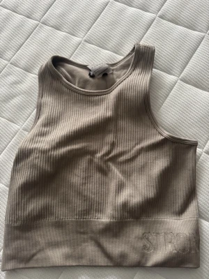 Beige ribbad linnetopp från Stronger - Snygg beige ribbad linnetopp från Stronger med broderad logga längst ner. Toppen är ärmlös och har en croppad passform som passar perfekt till höga byxor eller shorts. Materialet är mjukt och stretchigt, vilket gör den superbekväm. Rätt så liten i storleken