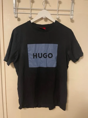  HUGO t-shirt - Snygg svart t-shirt från HUGO med ett stort blått tryck på bröstet där det står 'HUGO' i svart text. Klassisk rund halsringning och korta ärmar. Tillverkad i mjuk bomull som känns skön mot huden. Perfekt för en clean och modern look.