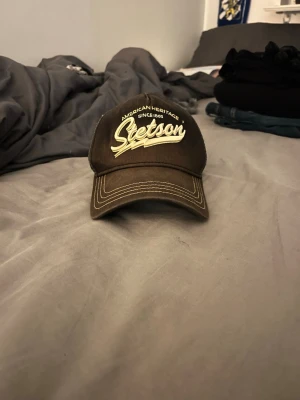Brun Stetson keps med brodyr - Säljer en brun keps från Stetson med broderad text 'American Heritage Since 1865' och stor logga framtill. Kepsen har böjd skärm och kontrastsömmar i beige. Perfekt för dig som gillar klassisk streetwear och vill ha en tidlös accessoar.
