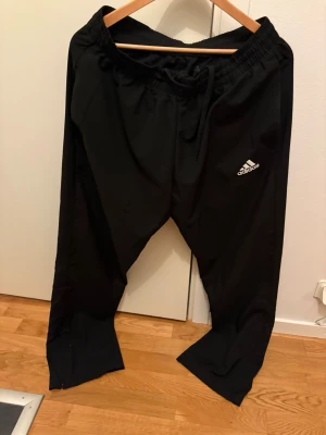 Svarta mjukisbyxor från Adidas - Svarta mjukisbyxor från Adidas med klassisk logga på låret. Byxorna har resår och dragsko i midjan för skön passform. Tillverkade i Primegreen-material som är återvunnet. Perfekta för chill eller träning.