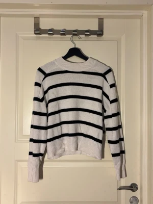 Vit stickad tröja med svarta ränder - Snygg vit stickad tröja med svarta horisontella ränder. Tröjan har rund halsringning, långa ärmar och ribbade muddar vid ärmslut och nederkant. Perfekt för en avslappnad och trendig look.