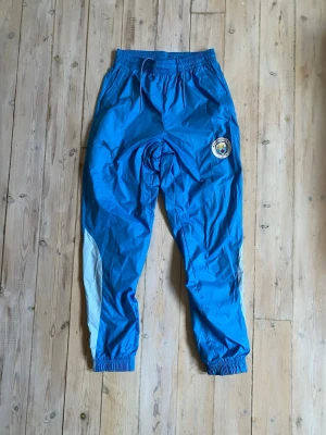 Blå Manchester City träningsbyxor Puma - Säljer ett par blå träningsbyxor från Puma med Manchester City-logga på ena benet. Byxorna har resår i midjan och vid bensluten samt vita detaljer längs sidorna. Perfekta för fotbollsträning eller chill. Tillverkade i lätt och snabbtorkande material.