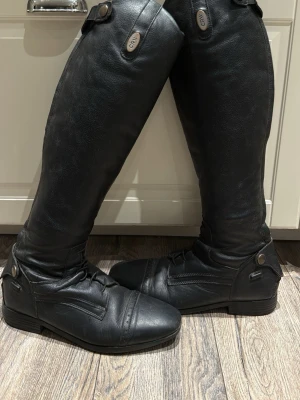 Svarta ridstövlar i läder från CRW - Säljer ett par svarta ridstövlar från CRW i äkta läder. Stövlarna har högt skaft, dragkedja på sidan och dekorativa knappar upptill. Klassisk design med låg klack och förstärkt tåparti. Perfekta för dig som gillar ridning och vill ha en stilren look.
