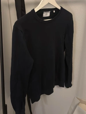 Mörkblå stickad tröja från Bläck - Stickad tröja från Bläck. Ribbstickade muddar vid hals, ärmslut och nederkant. Lite solblekt. Den är tillverkad i pimabomull och är i storlek S. 