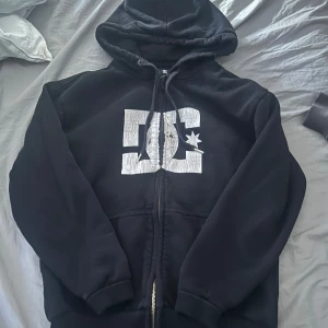 Svart DC hoodie med dragkedja - Svart hoodie från DC med stort vitt logotryck på bröstet. Tröjan har huva med snörning, dragkedja framtill och ribbade muddar vid ärmslut och nederkant. Perfekt för en avslappnad streetstil.