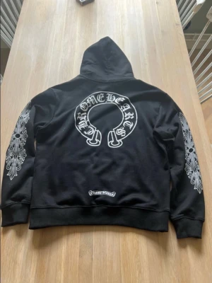 Chrome Hearts svart hoodie med tryck - Svart hoodie från Chrome Hearts med stora vita tryck på ryggen och ärmarna samt mindre logga på bröstet. Klassisk huva med dragsko och magficka framtill. Materialet är mjukt och bekvämt, perfekt för en streetwear-look.