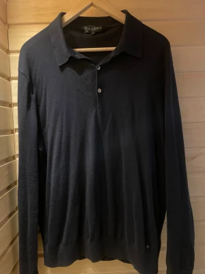 Svart långärmad tröja från Lagency - Klassisk svart långärmad tröja från Lagency med krage och två knappar framtill. Tröjan har en stilren design och är tillverkad i ett mjukt, tunt material som passar perfekt till många olika outfits.