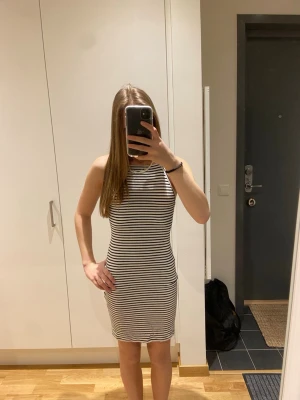 Randig vit och svart kort klänning - Säljer en tight, ärmlös kort klänning med smala axelband och rund halsringning. Klänningen är vit med svarta horisontella ränder och har öppen rygg. Perfekt för varma dagar och enkel att styla.