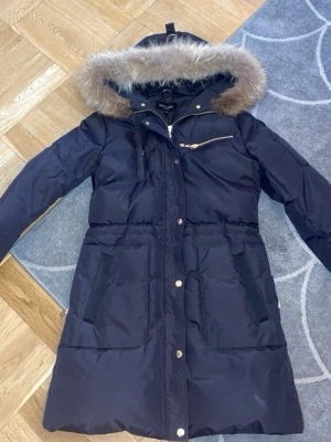 Hollies jacka med päls - Snygg svart parkas från Hollies med avtagbar huva och päls. Jackan har dragkedja och knappar framtill, flera fickor och en gulddetaljerad bröstficka. Perfekt för kalla dagar och riktigt stilren look.