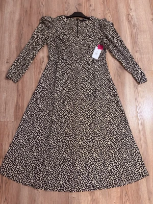 Elegant långklänning från Amelie & Me - Stilfull och tidlös klänning i storlek 42, helt ny och aldrig använd. Klänningen har ett snyggt prickigt mönster i svart och beige samt långa ärmar. Den smickrande passformen och A-linjeformade silhuetten ger ett feminint och elegant fall.  Perfekt både till vardags med boots eller sneakers och till festligare tillfällen med klackar och accessoarer. Ett säkert val för garderoben! 