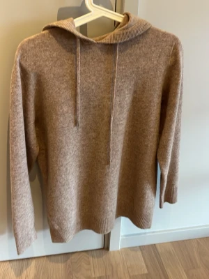 Beige stickad hoodie - Mysig beige stickad hoodie med huva och långa ärmar. Tröjan har ribbade muddar vid ärmslut och nederkant samt dragsko i huvan. Perfekt för en avslappnad och stilren look. Endast används en gång. Skriv vid funderingar!