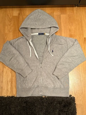 Grå zip-hoodie från Polo Ralph Lauren - Snygg grå hoodie med dragkedja från Polo Ralph Lauren. Klassisk design med huva och vita snören, samt den ikoniska lilla logon broderad på bröstet. Mjukt material och ribbade muddar vid ärmslut och nederkant. Perfekt för chill dagar. Storlek M passar S
