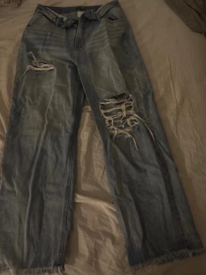 Ljusblå slitna raka jeans - Trendiga ljusblå jeans med raka ben och stora slitningar på båda benen. Jeansen har fransade benslut och klassisk femficksdesign. Perfekta för en avslappnad streetstyle-look.