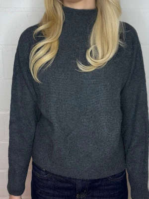 Mörkgrå stickad tröja med hög hals - Mysig mörkgrå stickad tröja med hög hals och långa ärmar. Tröjan har en rak passform och är tillverkad i ett mjukt, tjockare material som passar perfekt för kyligare dagar. Enkel och stilren design utan synliga loggor eller mönster.