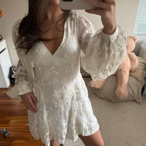 Vit blommig kortklänning med öppen rygg - Superfin vit kortklänning med blommigt mönster och volangdetaljer. Klänningen har v-ringning, långa puffärmar och öppen rygg med knyt. Perfekt för sommarens alla tillfällen som studenten eller kanske midsommar. 