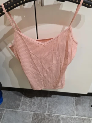 Ljusrosa ribbad linne - Säljer ett ljusrosa ribbat linne med smala axelband och v-ringning. Toppen har ett smickrande snitt och är perfekt för varma dagar. Materialet känns mjukt och stretchigt mot huden.