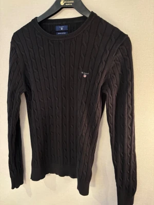 Svart kabelstickad tröja från GANT - Kabelstickad svart tröja från GANT i stretchig bomull. Tröjan har rund halsringning, ribbade muddar och GANT-logga broderad på bröstet. Perfekt för en stilren och klassisk look. Inga skador skick 7,5/10!🤝 storlek m men passar mer som S 