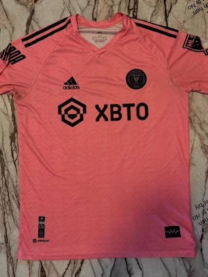 Inter Miami Messi matchtröja rosa - Snygg rosa matchtröja från Inter Miami med Messi och nummer 10 på ryggen. Tröjan har svarta detaljer, klubbmärke och Adidas-logga framtill samt sponsortryck. Tillverkad i lätt och ventilerande material, perfekt för fotboll eller träning.