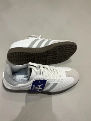 Samba sneakers - Samba sneakers i vitt med gråa detaljer och de tre ränderna på sidan. Ovandelen är i läder med mockadetaljer framtill och yttersulan är i gummi. Perfekta för dig som gillar retrostil och streetwear.