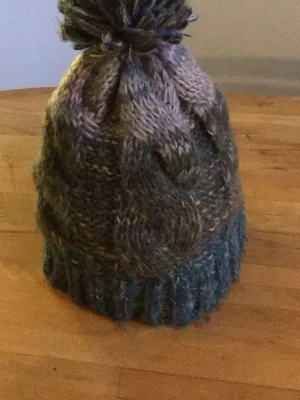 Stickad mössa med tofs i blågrå toner - Mysig stickad mössa i blågrå och beige melerad ull med stor tofs på toppen. Mössan har ett flätstickat mönster och bred ribbad kant nedtill. Perfekt för kalla dagar och ger en avslappnad, trendig look.