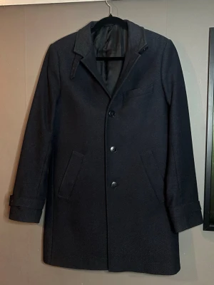 Marinblå  klassisk ullrock med knappar - Stilren marinblå rock från Riley i ullblandning med tre knappar framtill, snedställda fickor och enkel krage. Rocken har slät insida och justerbara spännen vid ärmsluten. Perfekt för kyliga dagar och ger en tidlös look.