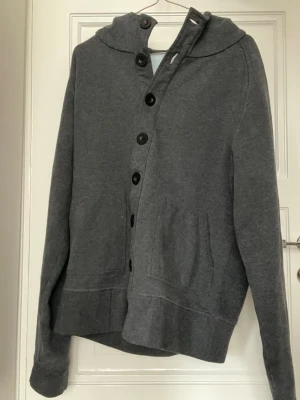 NN07 hoodie  - Mörkgrå hoodie från NN07 med huva och stora knappar framtill. Tillverkad i mjukt sweatshirtmaterial och har två fickor på magen. Perfekt för en avslappnad och chill stil. Passar dig som gillar enkel och stilren design.