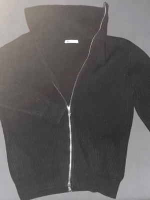 Svart ribbad ziptröja från Zara - Snygg svart ribbad ziptröja från Zara med hög krage och hel dragkedja framtill. Tröjan har långa ärmar och är tillverkad i ett mjukt stickat material som ger en clean och stilren look. Perfekt att slänga på sig över en t-shirt.