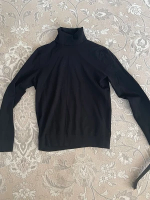 Svart polotröja i stickat material - Säljer en svart stickad polotröja med lång ärm och klassisk passform. Tröjan har hög polokrage och är tillverkad i ett mjukt, tunt material som passar perfekt till lager på lager. Enkel och stilren design utan synliga detaljer eller mönster.