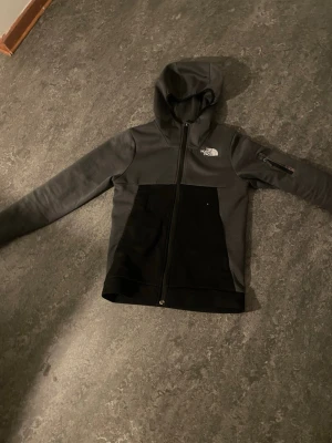Svart och grå hoodie från The North Face - Snygg hoodie från The North Face i svart och mörkgrått. Modellen har dragkedja framtill, huva och en praktisk ficka med dragkedja på ärmen. Perfekt för en sportig och avslappnad stil. Materialet är mjukt och bekvämt, passar till många tillfällen.