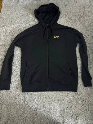 Svart EA7 hoodie med dragkedja - Svart hoodie från EA7 med dragkedja framtill och två fickor. Liten gul EA7-logga på bröstet. Klassisk huva med dragsko och ribbade muddar. Perfekt för en chill och stilren look.
