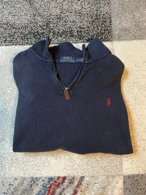 Mörkblå halvzip från Polo Ralph Lauren - Mörkblå långärmad tröja från Polo Ralph Lauren med halvzip och klassisk krage. Tröjan har röd broderad logga på bröstet och ribbade muddar vid ärmslut och nederkant. Perfekt för en stilren och avslappnad look.