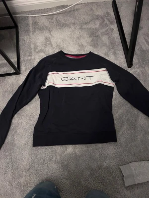 Mörkblå sweatshirt från GANT - Snygg mörkblå sweatshirt från GANT med vit panel och röd detalj över bröstet samt stor logga framtill. Tröjan har rund halsringning och långa ärmar. Perfekt för en avslappnad och trendig stil.säljer den för den har blivit för liten 12-11 år skulle ja säga den passar. Pris kan diskuteras Mvh Felix⭐️