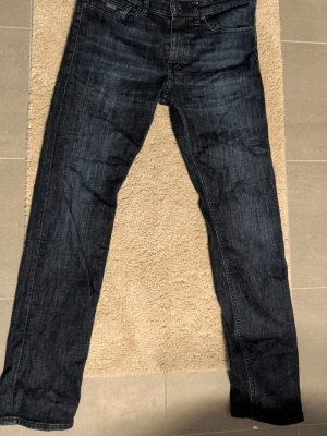 Mörkblå Boss Delaware Slim Jeans - Säljer ett par mörkblå jeans från BOSS, modell Delaware Slim Fit. Jeansen har klassisk femficksdesign, raka ben och snygg tvättad finish. Märkespatch i läder baktill i midjan och diskreta sömmar. Perfekta för dig som gillar stilrena och moderna jeans.