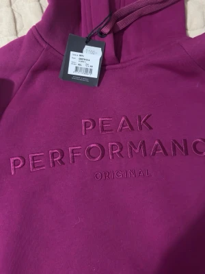 Lila hoodie från Peak Performance - Snygg lila hoodie från Peak Performance med broderad logga på bröstet. Klassisk huva med dragsko och stor magficka framtill. Tillverkad i mjukt bomullsmaterial och har ribbade muddar vid ärmslut och nederkant. Perfekt för en avslappnad och sportig stil.