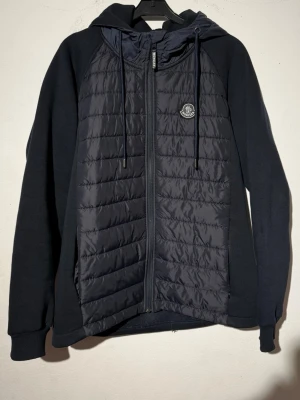 Mörkblå vindjacka från Moncler - Snygg mörkblå vindjacka från Moncler med quiltad front, dragkedja och huva med dragsko. Jackan har Moncler-logga på bröstet och mjuka ärmar i sweatshirtmaterial. Perfekt för dig som vill ha en sportig och stilren look.