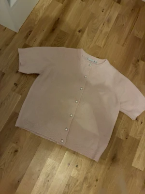 Ljusrosa kofta med pärlknappar - Supersöt ljusrosa kofta med korta ärmar och pärlknappar framtill. Rak och lite boxig passform, rund halsringning och ribbade kanter. Perfekt för att addera en mjuk och feminin touch till din outfit.