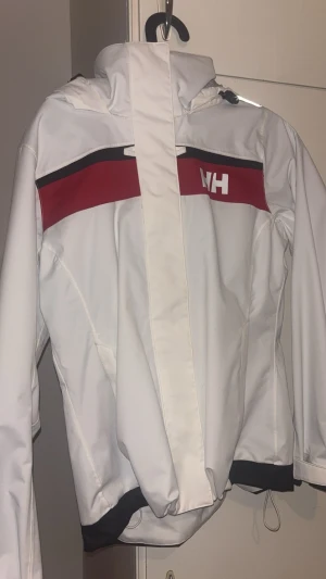 Vit vindjacka från Helly Hansen - Snygg vit vindjacka från Helly Hansen med röd och svart detalj över bröstet och HH-logga. Jackan har huva, dragkedja och justerbar nederkant. Perfekt för kalla och blåsiga dagar. Materialet är vattenavvisande och fodret har meshpaneler. Använd men inget tecken på slitage, säljer för kommer inte till användning så legat i en garderob i några år.