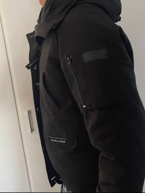 Svart dunjacka från Canada Goose - Säljer en svart dunjacka från Canada Goose med huva och flera fickor, inklusive en praktisk ficka med dragkedja på ärmen. Jackan har en stilren design och är perfekt för kalla vinterdagar. Klassisk logga på fickan och robust dragkedja framtill.passar st/M