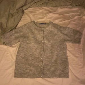 Ljusgrå kofta med kort ärm - Mysig ljusgrå kofta med korta ärmar och rund hals. Koftan har knappar framtill och är stickad i ett mjukt material som ger en avslappnad look. Perfekt att slänga över en t-shirt eller linne för extra lager. Storlek XS/34