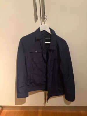 Mörkblå vindjacka från Lardini - Stilren mörkblå vindjacka från Lardini med klassisk krage, dragkedja och tryckknappar framtill. Jackan har två bröstfickor med lock och raka ärmar. Perfekt för dig som vill ha en enkel och snygg look.