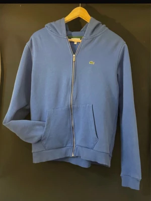 Blå zip-hoodie från Lacoste - Snygg blå hoodie med dragkedja från Lacoste. Klassisk krokodillogga på bröstet, huva och stora fickor framtill. Perfekt för en avslappnad och sportig stil. Mjuk bomullskänsla och ribbade muddar vid ärmslut och nederkant.