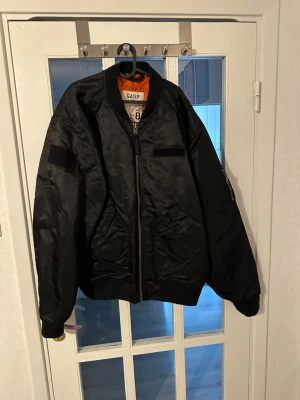 Svart bomberjacka från GASP - Cool svart bomberjacka från GASP med orange foder, klassisk passform och dragkedja framtill. Jackan har stora fickor, ribbade muddar och är tillverkad i glansigt syntetmaterial. Perfekt för dig som gillar streetwear och sportig stil.