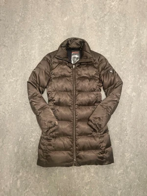 Prada Lång brun jacka - Snygg brun lång dunjacka från Prada med quiltad design och dragkedja framtill. Jackan har hög krage och diskret logotyp på ärmen. Perfekt för kalla vinterdagar och ger en stilren look. Model nummer är Art.29C150, Skriv gärna om det är något😁