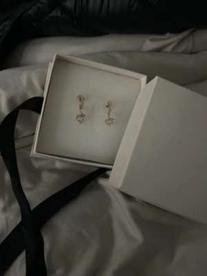 Guldiga örhängen med hjärtan och stenar - Säljer ett par guldiga örhängen med små gnistrande stenar. Örhängena har en hängande hjärtformad detalj som är täckt av klara stenar för extra bling. Perfekta för dig som vill ha något sött och stilrent med lite extra shine.