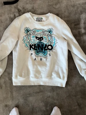 Vit Kenzo sweatshirt med tigertryck - Vit sweatshirt från Kenzo med ikoniskt broderat tigerhuvud i blått och svart samt texten 'KENZO PARIS' framtill. Tröjan har rund halsringning och ribbade muddar vid ärmslut och nederkant. Perfekt statement-plagg för dig som gillar streetwear.
