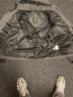 Svart dunjacka från Canada Goose - Säljer en svart dunjacka från Canada Goose, modell 4600M. Jackan har en klassisk design med slät yta och svart innerfoder. Perfekt för kalla vinterdagar tack vare den rejäla dunfyllningen. Märkeslogga finns på insidan.