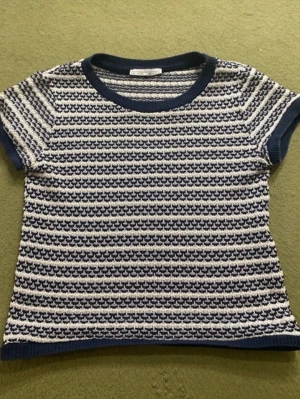 Randig stickad topp i blått och vitt - Snygg kortärmad topp med blå och vita ränder i ett stickat material. Rund halsringning och ribbade kanter vid ärmar, hals och nederkant. Perfekt för dig som gillar retrovibbar och vill ha något bekvämt men ändå stilrent.