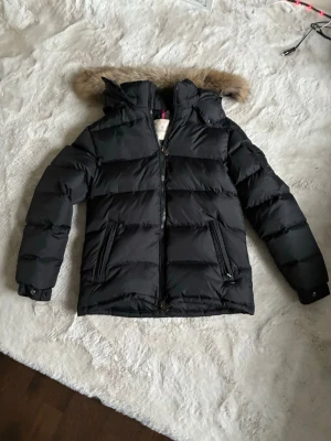 Svart Moncler pufferjacka med päls  - Svart pufferjacka från Moncler med huva och snygg pälsdetalj men kan ta av pälsen också. Jackan har dragkedja framtill, fickor med dragkedja och en Moncler-logga på ärmen. Materialet är polyester och dun, perfekt för kalla dagar. Stilsäker och varm jacka med normal passform. En helt ny jacka använd en gång 