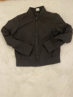 Svart bomberjacka Urban Classics - Snygg svart bomberjacka från Urban Classics med dragkedja framtill och ribbade muddar vid ärmslut och midja. Jackan har klassisk bombermodell och är tillverkad i ett lätt, slätt material. Perfekt för en stilren och urban look.